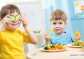 Zwei Jungs im Kitaalter beim gemeinsamen Essen. auf dem Tisch stehen Teller mit buntem Gemüse. Ein Junge hält sich zwei Gurkenscheiben vor die Augen