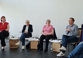 drei Frauen und ein Mann sitzen auf dem Podium, rechts flankiert von einem Roll-up mit der Aufschrift "Aufstehen für Demokratie und Menschenrechte"